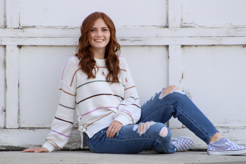 A young woman with long, wavy auburn hair and a friendly smile sits on a concrete ledge. She is wearing a white oversized sweater with thin, multi-colored horizontal stripes and lace-up details on the sides, paired with distressed blue jeans and checkered slip-on sneakers. She is posed in front of a weathered, white-paneled garage door.