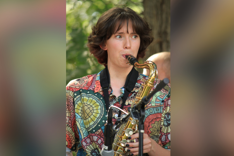 A person with short, wavy brown hair plays a brass saxophone during an outdoor performance. They are wearing a vibrant, multicolored button-down shirt with a bold circular and floral pattern. Their nails are painted black, and they are looking off to the side with focus while playing into a microphone. The background is filled with soft-focus green trees.