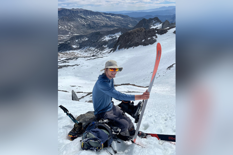 A young man sits on a rock atop a snow-covered mountain peak, smiling while holding up a single long ski. He is wearing a blue long-sleeved hooded shirt, grey hiking pants, a tan sun hat, and orange-tinted mirrored sunglasses. Next to him are a backpack, hiking boots, and trekking poles settled in the snow. The background reveals a vast, rugged mountain range under a blue sky with soft white cloud