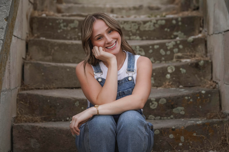 A young woman with shoulder-length light brown hair and a bright smile sits on a set of weathered stone steps. She is leaning her chin on her hand in a relaxed, thoughtful pose. She is wearing blue denim overalls over a white sleeveless top, accessorized with a gold watch and a delicate necklace. The steps behind her are aged with patches of moss and lichen.