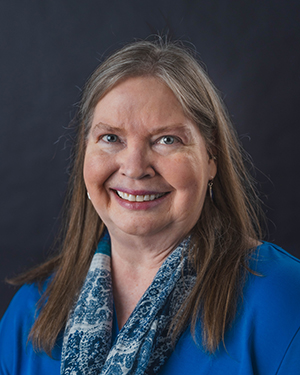 A close-up portrait of a mature woman with light brown and grey hair and blue eyes smiling warmly. She is wearing a bright blue top paired with a blue and white patterned scarf. The photo is taken against a solid dark grey studio backdrop with professional, even lighting.