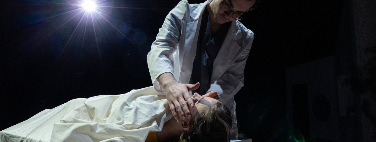 Actors on stage portray a doctor examining a dead body