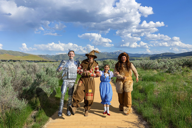Characters in the play Wizard of Oz including Dorothy, Lion, Tin Man and Scarecrow walk on a yellow brick road with green grass around them and blue skies above them.