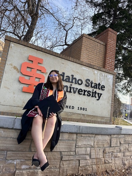 A graduate sits by an Idaho State University sign